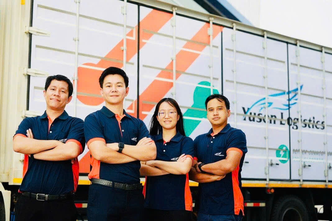 Nhân viên Yusen Logistics mặc áo polo đồng phục xanh cam trước xe vận tải.