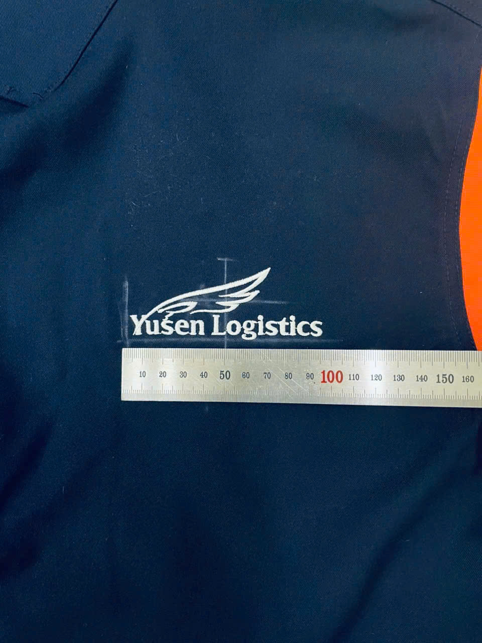 Kiểm tra kích thước logo thêu Yusen Logistics trên vải áo bằng thước.