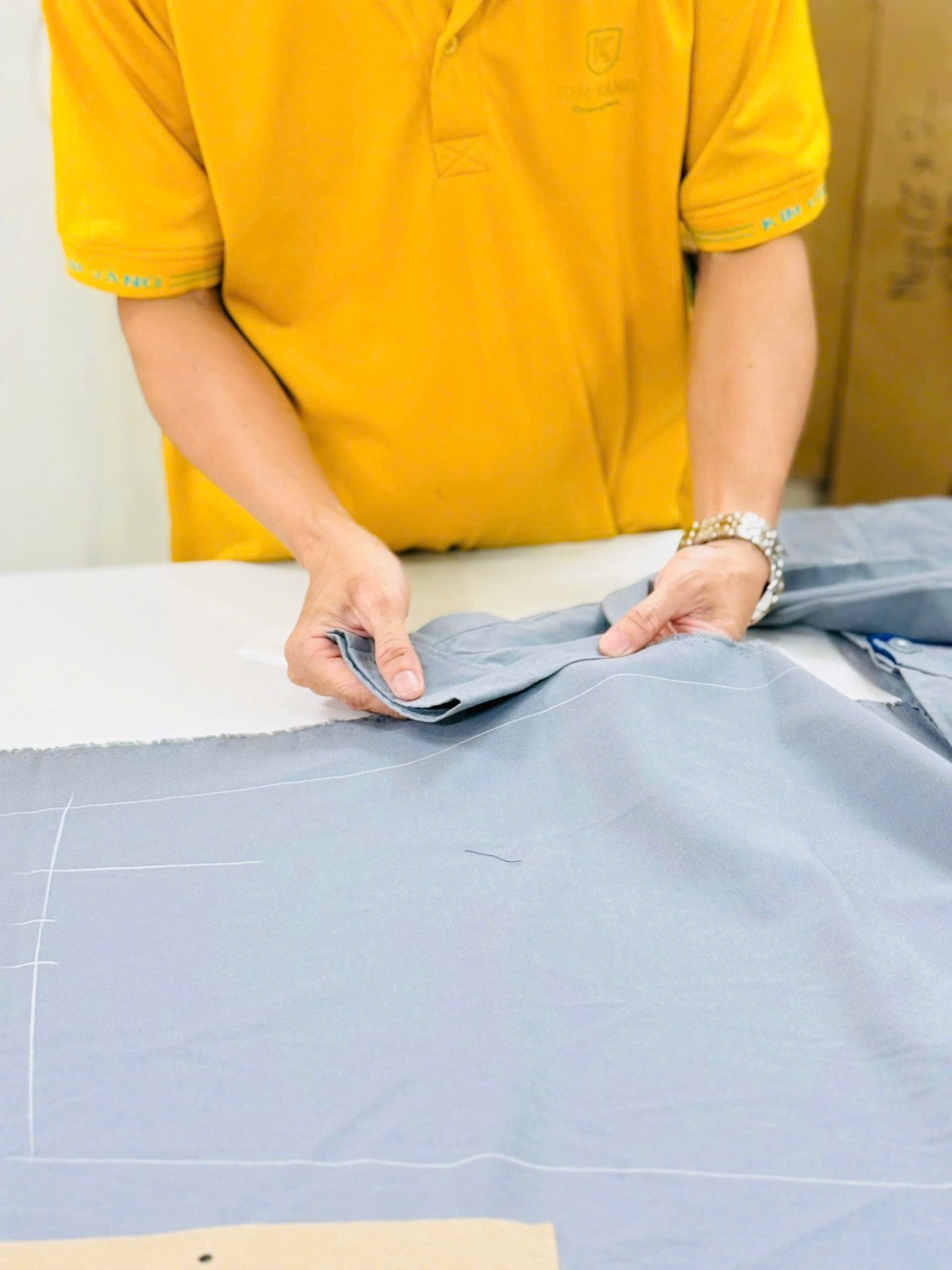 Bàn tay người thợ đối chiếu mép chi tiết vải xám trên bàn kỹ thuật.